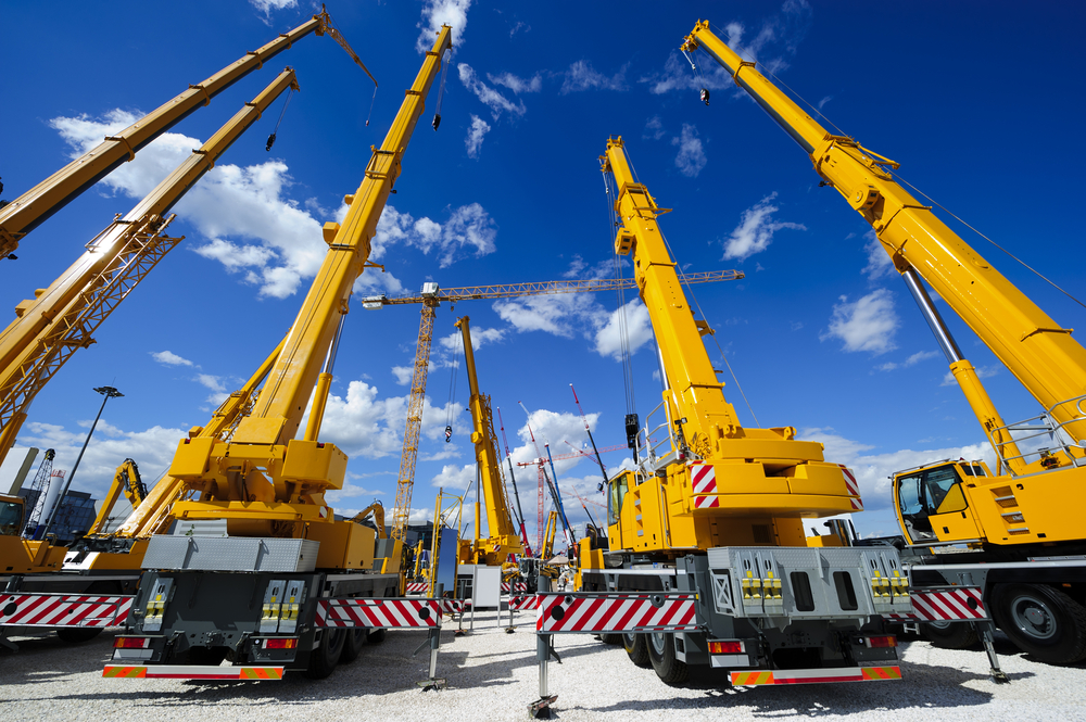Mobile-construction-cranes-with-yellow-telescopic-arms-and-big-tower-cranes-in-sunny-day-with-white-clouds-and-deep-blue-sky-on-background