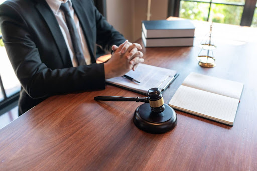 Man in a suit seated at a desk with hands clasped, beside a gavel, legal documents, open notebook, and stacked books, with sunlight coming through nearby windows. do personal injury lawyers always win